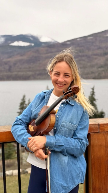Marta holding her violin outdoors with mountains and a lake in the background